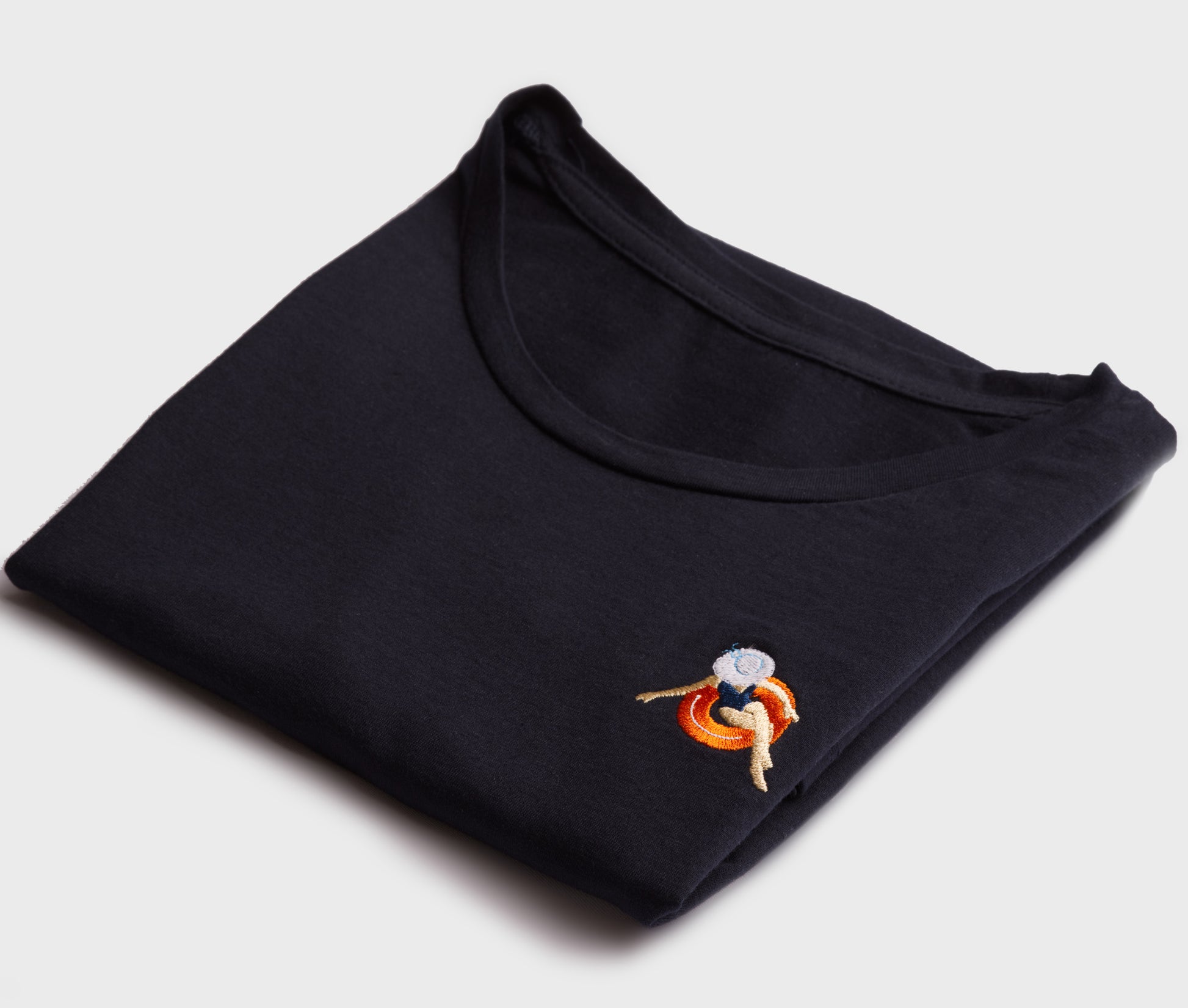 A dark navy blue crewneck t-shirt folded neatly on a light background, highlighting an embroidered patch on the chest featuring a person lounging on an orange pool float.